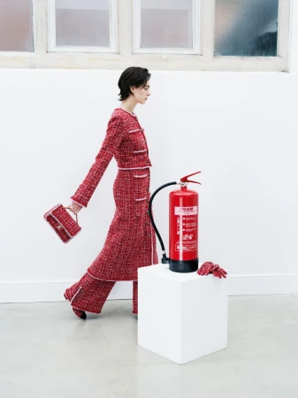 Woman in a red tweed suit walks past a red fire extinguisher on a white block, against a white wall and window background.