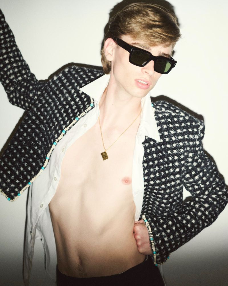 A young adult male poses in a studio wearing a patterned jacket, open shirt, and black sunglasses, showcasing a fashion-forward style.