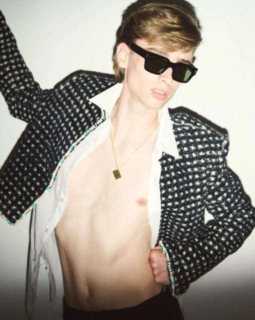 A young adult male poses in a studio wearing a patterned jacket, open shirt, and black sunglasses, showcasing a fashion-forward style.