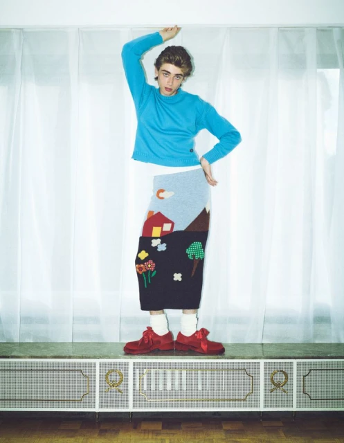 Person in a turquoise sweater and graphic skirt stands indoors against a white curtain and paneled wall, posing with arms raised.