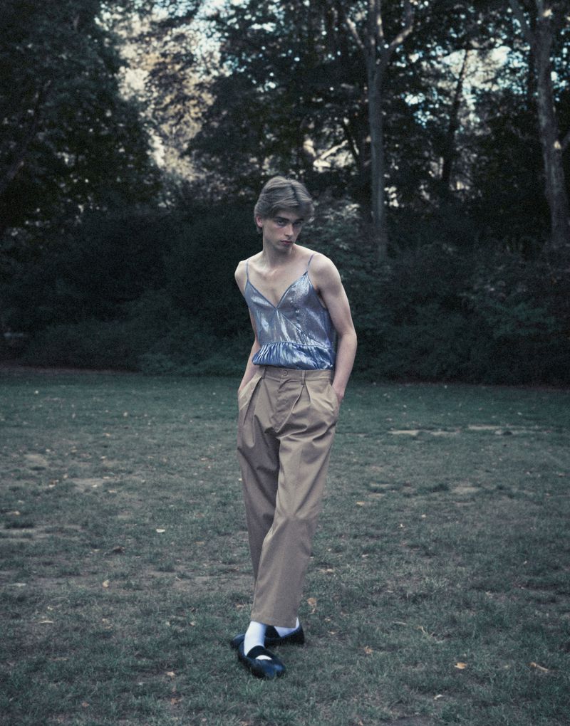 Person in a shiny blue top and khaki pants stands on a grassy lawn surrounded by trees. Fashion editorial on an outdoor set.