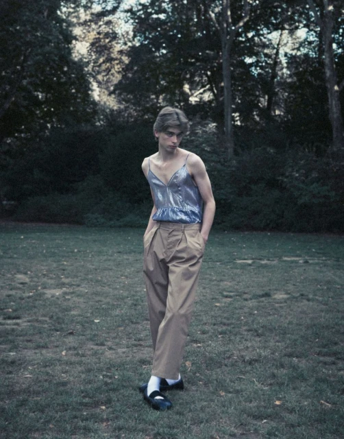 Person in a shiny blue top and khaki pants stands on a grassy lawn surrounded by trees. Fashion editorial on an outdoor set.