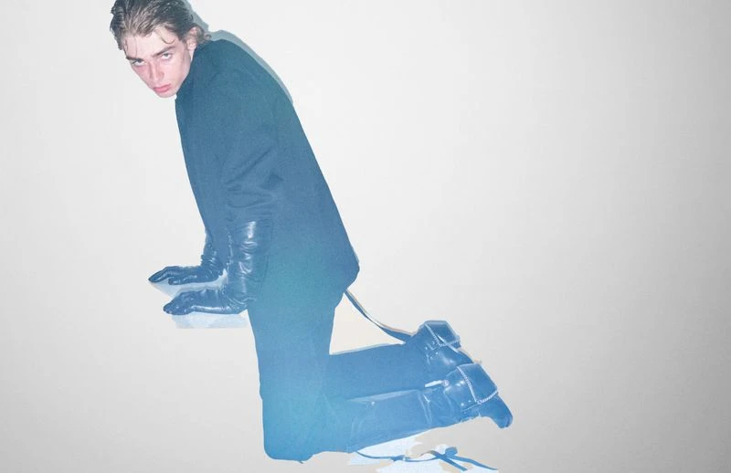 Male model in a black suit and leather gloves poses kneeling against a white wall in studio lighting for a fashion editorial photoshoot.