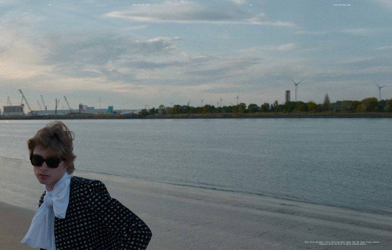 Adult male in sunglasses poses on a beach near a river with a city skyline, wind turbines, and overcast skies in the background.