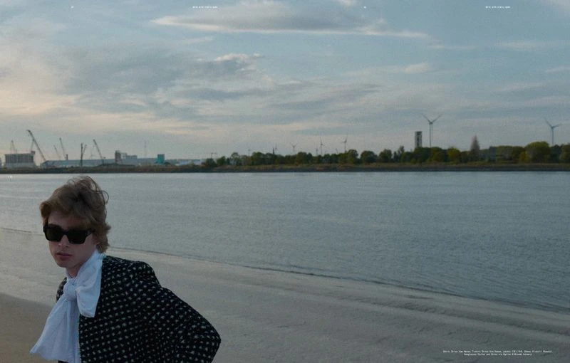 Adult male in sunglasses poses on a beach near a river with a city skyline, wind turbines, and overcast skies in the background.