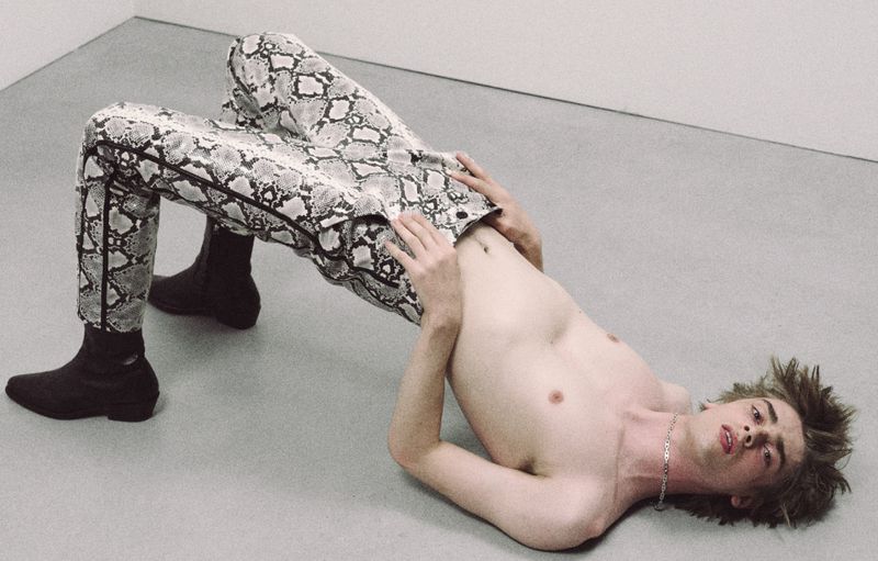 A shirtless young man wearing snake-patterned pants and boots poses in a bridge position against a neutral wall and floor background.