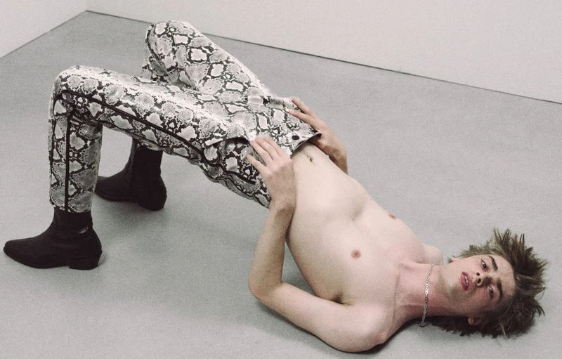 A shirtless young man wearing snake-patterned pants and boots poses in a bridge position against a neutral wall and floor background.