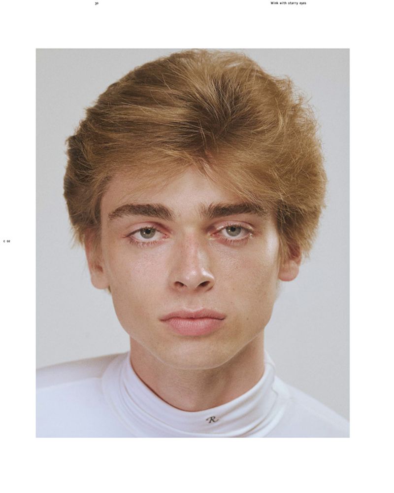 Close-up portrait of model Luka Hoogedeure with textured blonde-brown hair wearing a white mock-neck top against a light grey background.