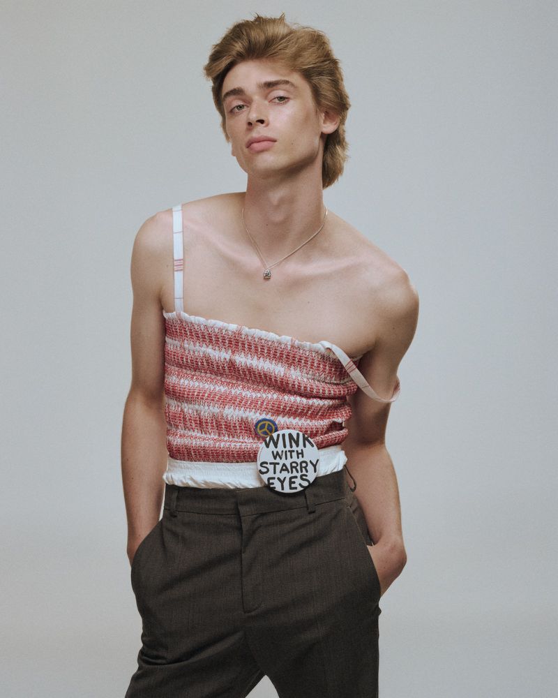 Young adult male poses indoors wearing a hand-crafted pink and white striped top and dark trousers against a plain light background.