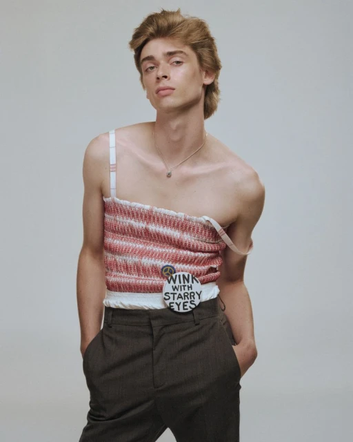Young adult male poses indoors wearing a hand-crafted pink and white striped top and dark trousers against a plain light background.