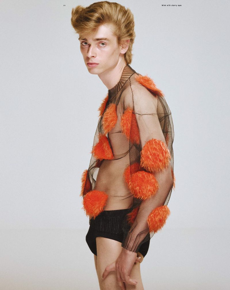 Male fashion model poses in sheer black shirt with orange tufts against a plain white background, showcasing unique editorial style.