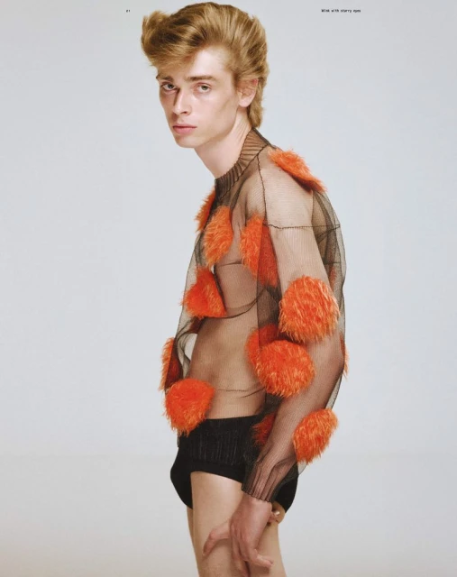 Male fashion model poses in sheer black shirt with orange tufts against a plain white background, showcasing unique editorial style.