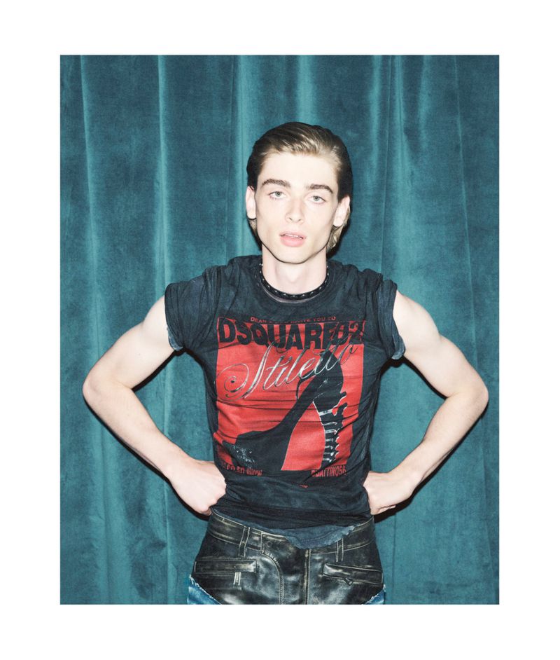 A young adult man poses in a dark graphic tee and leather-look pants against a turquoise curtain backdrop in a studio fashion shot.