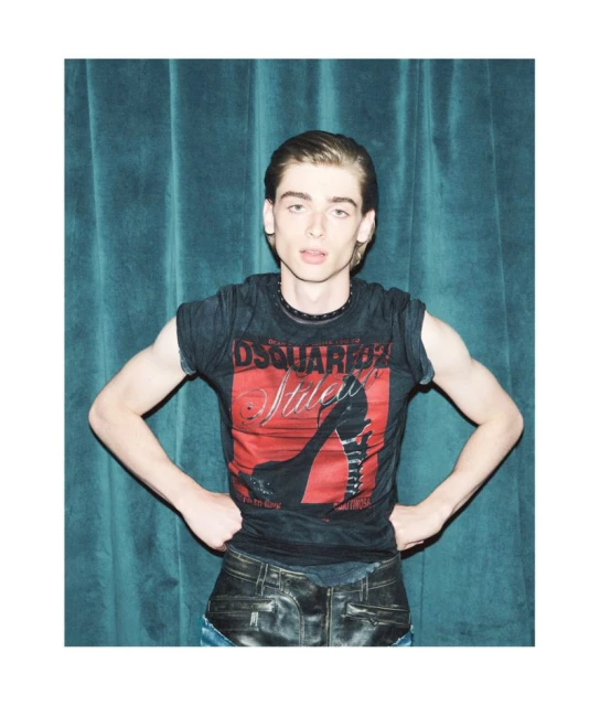 A young adult man poses in a dark graphic tee and leather-look pants against a turquoise curtain backdrop in a studio fashion shot.