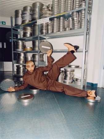Young adult man in brown jumpsuit poses creatively with silver film canisters in a storage room with stacked canisters on shelves.