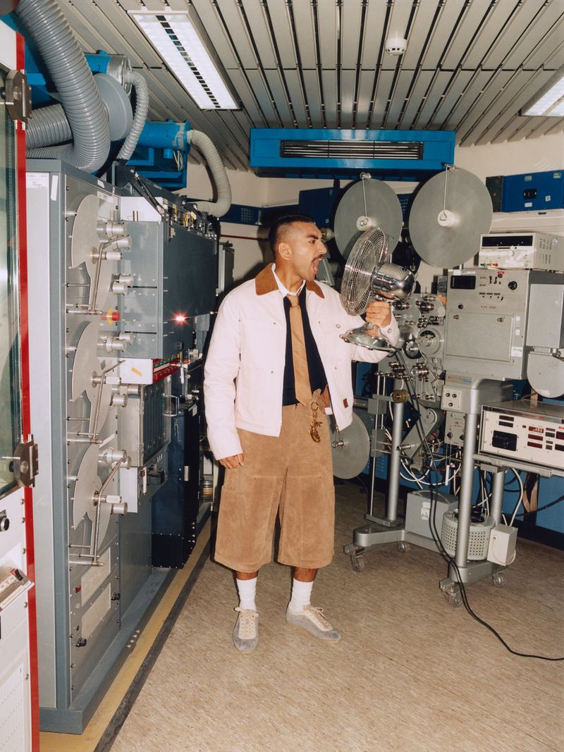 Man in beige jacket and shorts holds fan in an industrial setting with machines and equipment, facing left with a neutral expression.