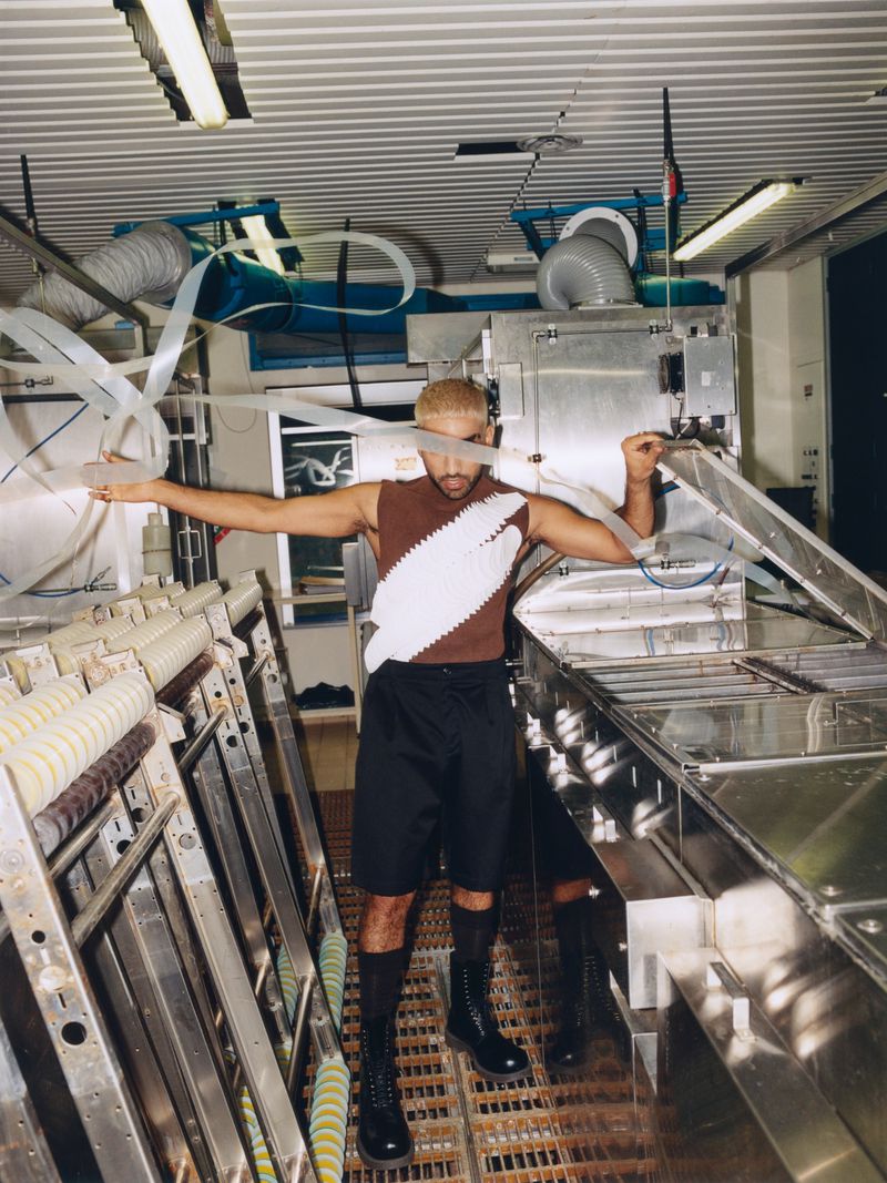 Adult male poses in an industrial setting, wearing a brown sleeveless shirt, black shorts, socks, and boots, surrounded by metal machinery.