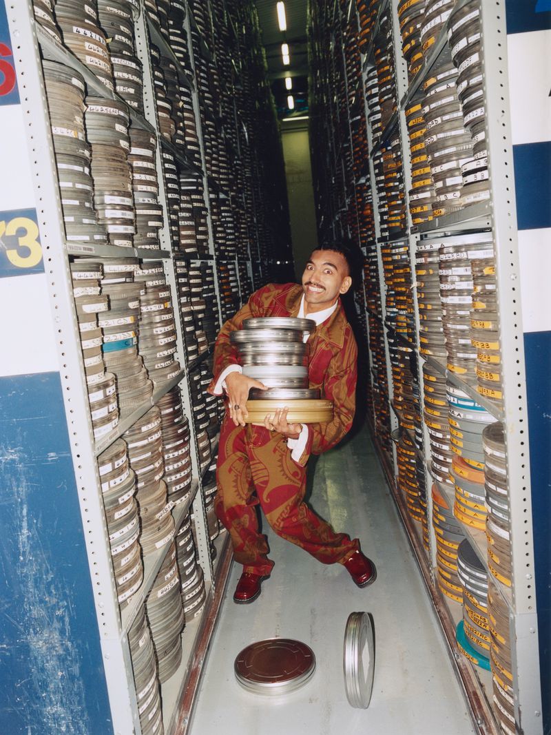 Man in patterned suit holds film reels in storage, surrounded by rows of film canisters on shelves. Storage with rows of film.