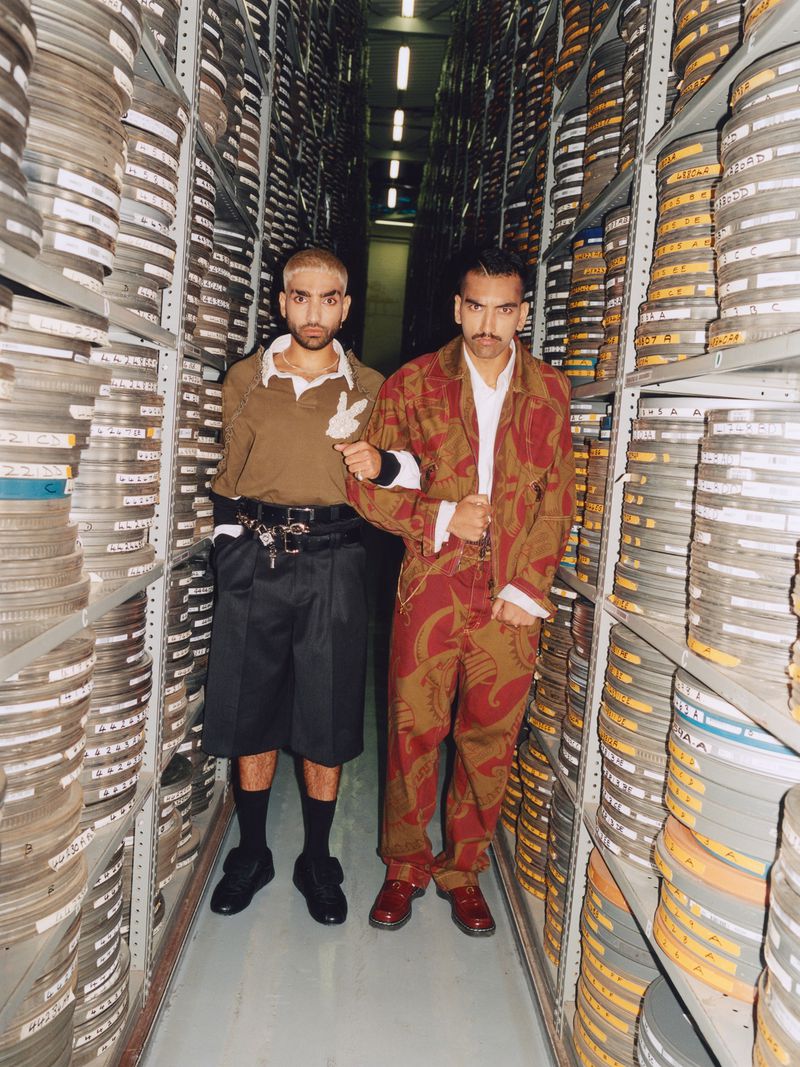 Two adult males pose indoors in a hallway filled with shelves containing reels of film, dressed in eye-catching patterned clothing.