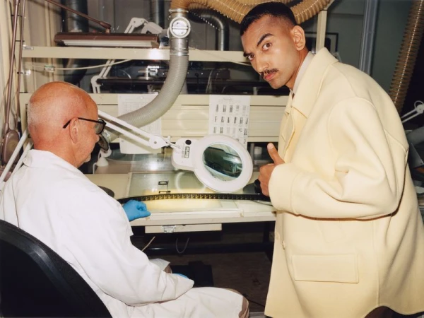 Film technician inspecting film strip with magnifying glass while man in coat looks on giving a thumbs up in laboratory setting.