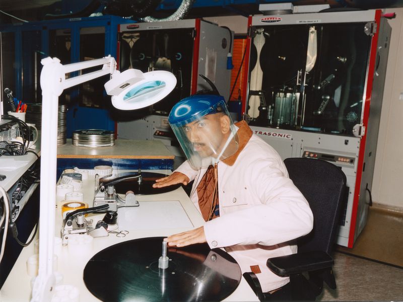 Man in cleanroom suit works with large black disk under bright light and magnifying lamp, in what appears to be a factory setting.