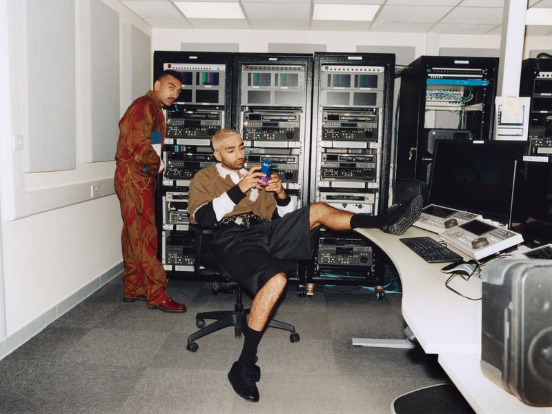 Two adult males pose indoors in a room filled with computer servers, one seated in an office chair using his cell phone.