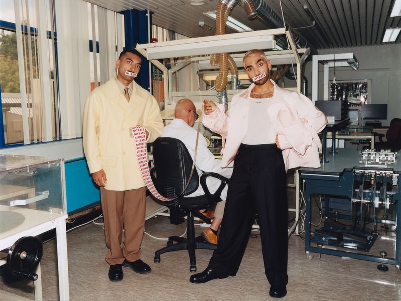 Two men in lab coats with 'Nitrate' tape over their mouths stand in a lab, another man sits at a desk in the background.