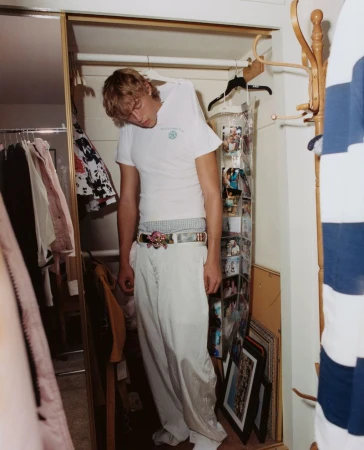 Young man with blonde hair in white t-shirt and loose pants standing inside of a closet with hanging photos and a coat rack.