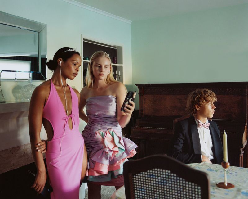 Two women in formal dresses, one using a smartphone, stand next to a young man in a tuxedo seated at a table indoors.