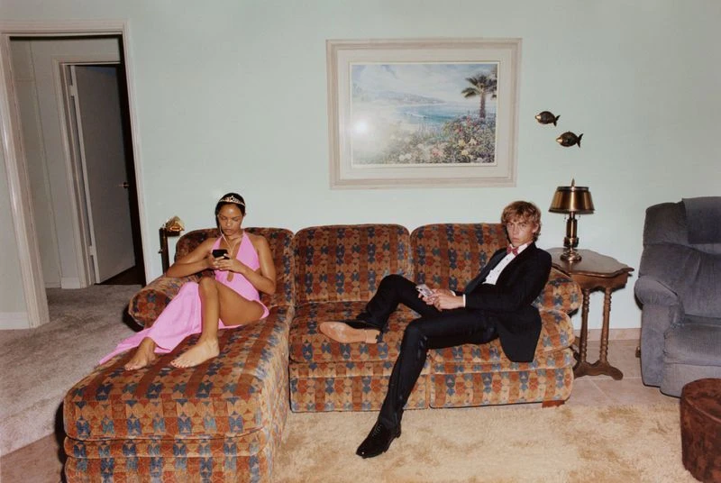 A barefoot couple lounges on a patterned couch in a retro-styled living room. The woman looks at her phone, while the man sits in a suit.