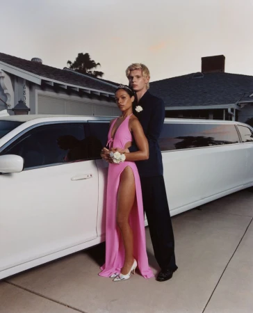 Young couple in formal attire posing in front of a white limousine and a house at dusk, the woman wears a pink gown.