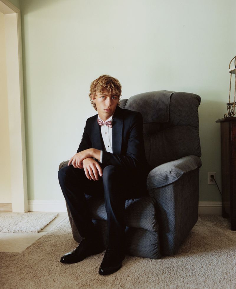 Blond male in a suit and bow tie poses while sitting in a gray armchair indoors against a pale green wall, looking at the camera.