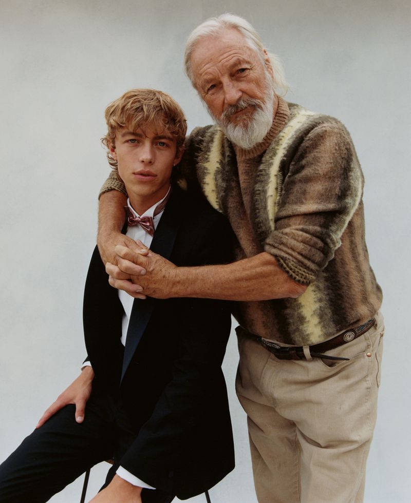 Indoor portrait of a young adult male in a black suit seated next to an older adult male with a white beard, both wearing formalwear.