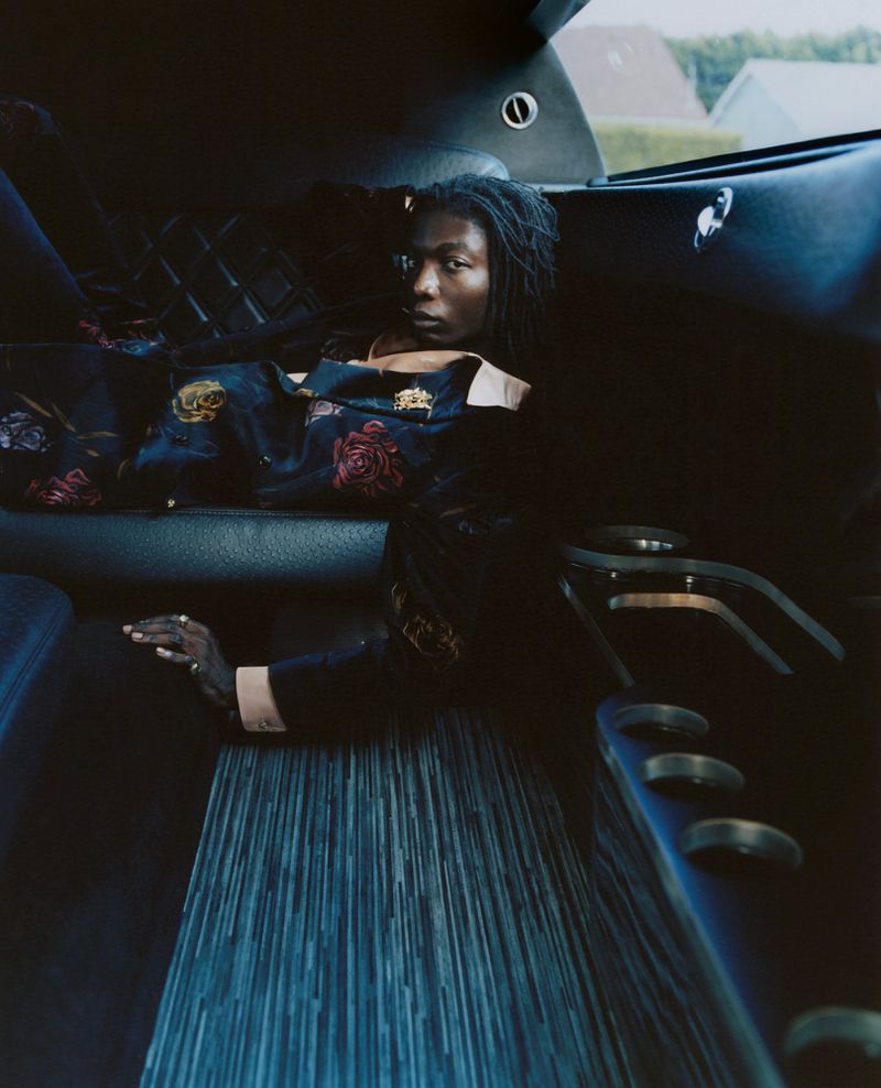 A Black woman with dreadlocks reclines on a blue striped seat inside a limo wearing a floral shirt, looking directly at the viewer.