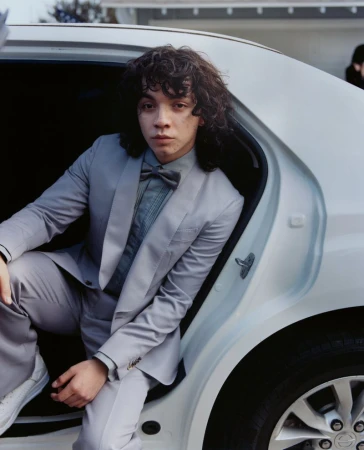 Young adult in a grey suit poses in a white car, featuring curly hair, a bow tie, and white sneakers, set against an urban backdrop.
