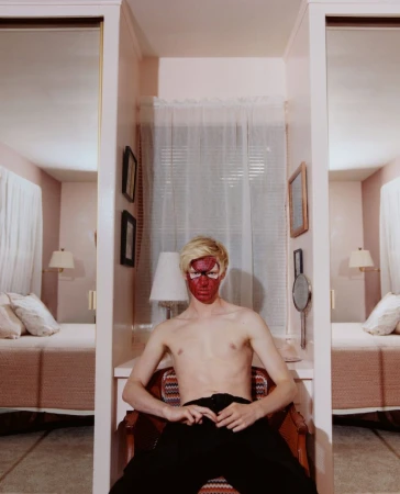 A person with red paint on their face poses while seated in a vintage chair in a pink-toned room featuring a bed, window, and mirror.