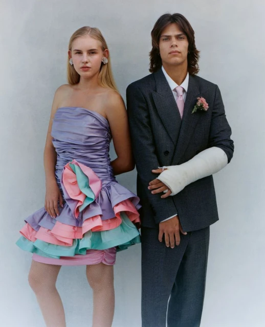 Two young people pose in formal wear against a white backdrop; the male has a cast on his arm; portrait in a photo studio.