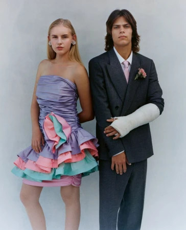 Two young people pose in formal wear against a white backdrop; the male has a cast on his arm; portrait in a photo studio.