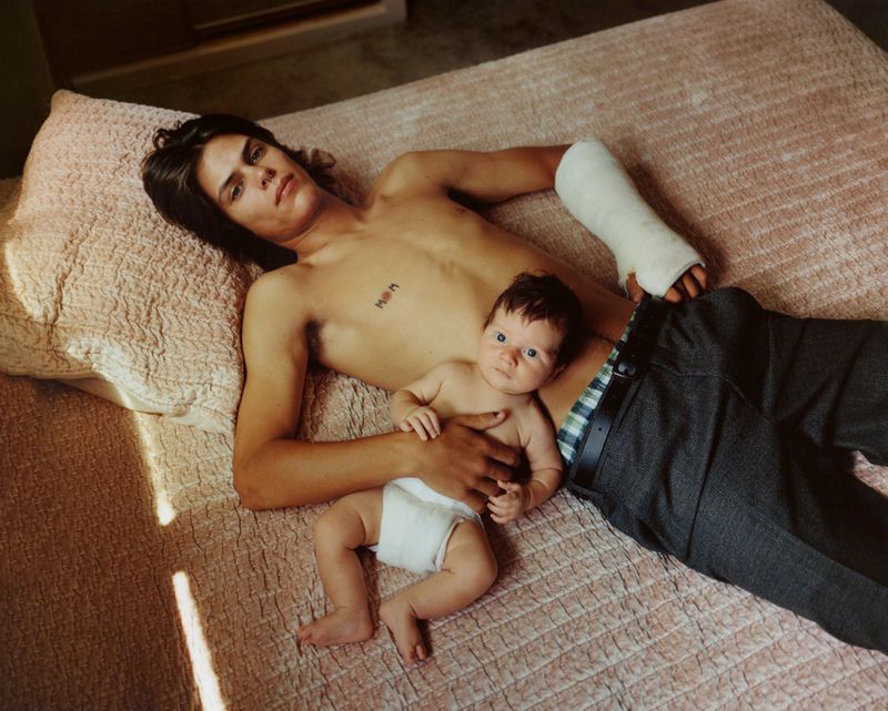 Adult male with cast on arm and baby lying on bed, medium shot, indoor setting, warm lighting, casual attire, portrait photography.