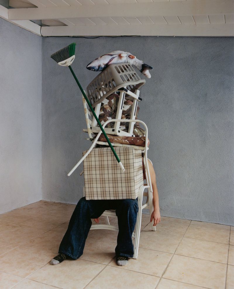 Person seated under a stack of household objects including a laundry basket, cushions, boxes, and a broom in an indoor environment.