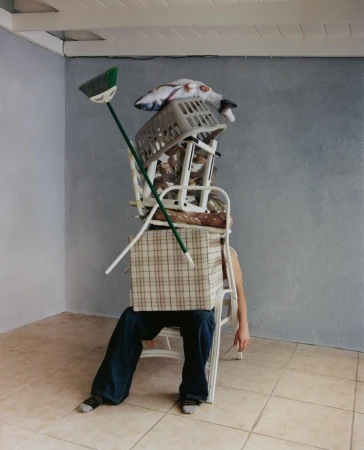 Person seated under a stack of household objects including a laundry basket, cushions, boxes, and a broom in an indoor environment.