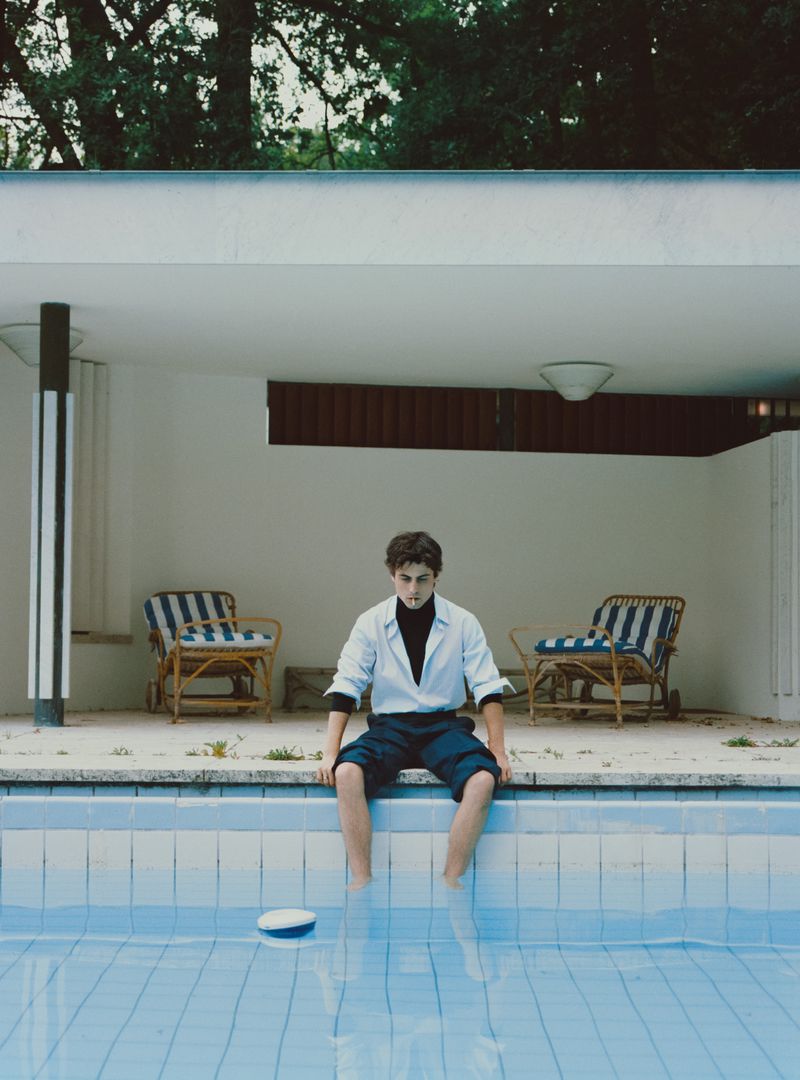 Young man in white shirt and dark shorts sits by the pool, with a white hat floating in the water. Chairs line the background.
