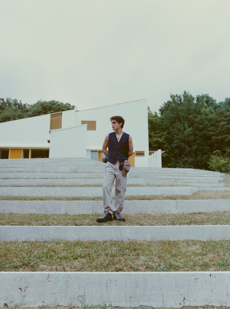 A young man descends concrete steps in a vest and khaki pants, with a modern white building and lush trees in the background.