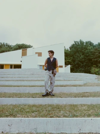 A young man descends concrete steps in a vest and khaki pants, with a modern white building and lush trees in the background.
