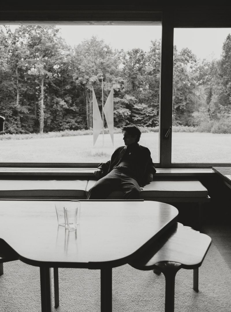 Monochrome portrait of a person sitting by a window with a view of trees, featuring a minimalist table and glass in the foreground.