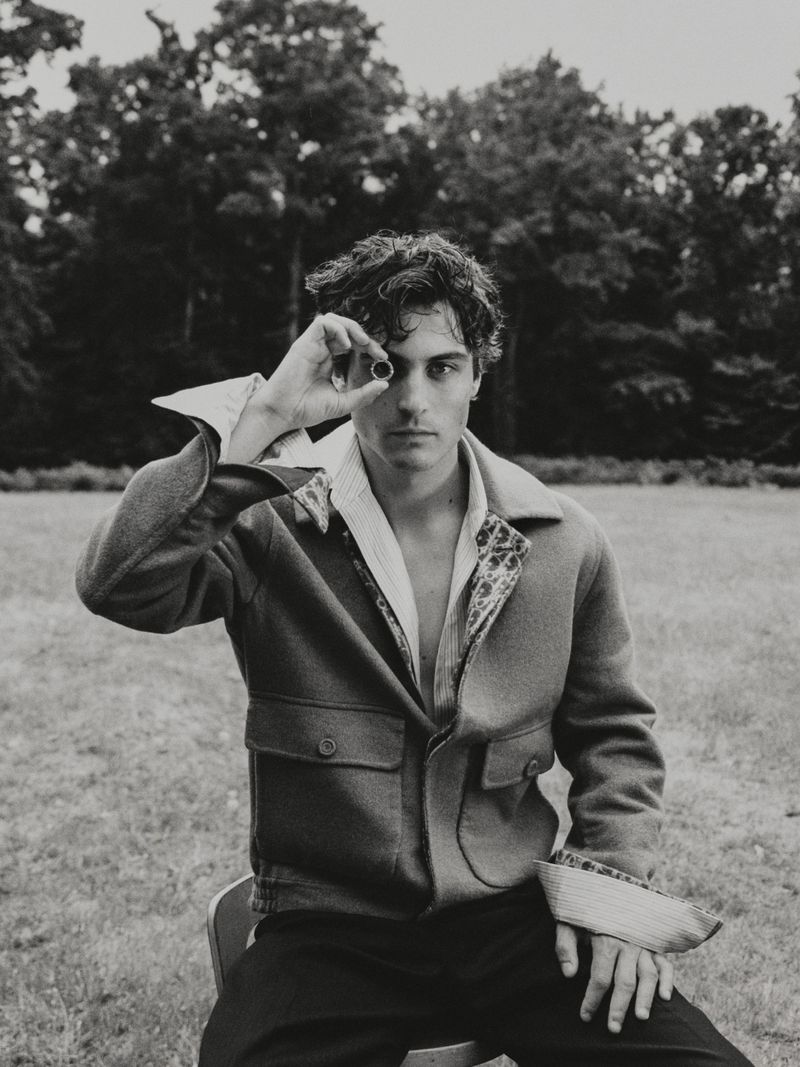Monochrome portrait of a young adult man sitting on a chair outdoors, holding a small, dark object to one eye in a grassy field.