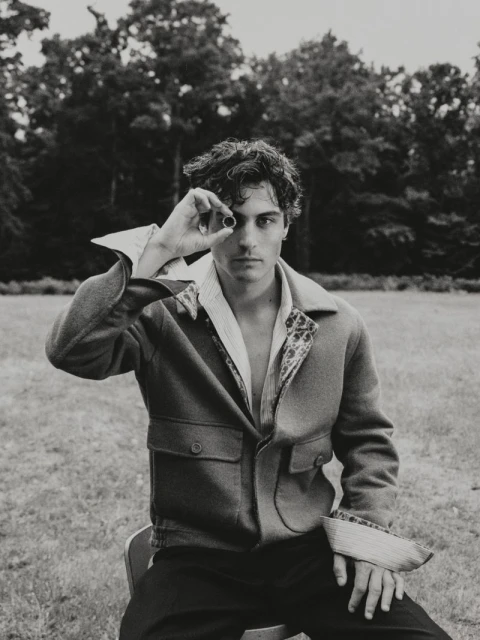 Monochrome portrait of a young adult man sitting on a chair outdoors, holding a small, dark object to one eye in a grassy field.