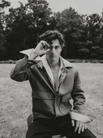 Monochrome portrait of a young adult man sitting on a chair outdoors, holding a small, dark object to one eye in a grassy field.