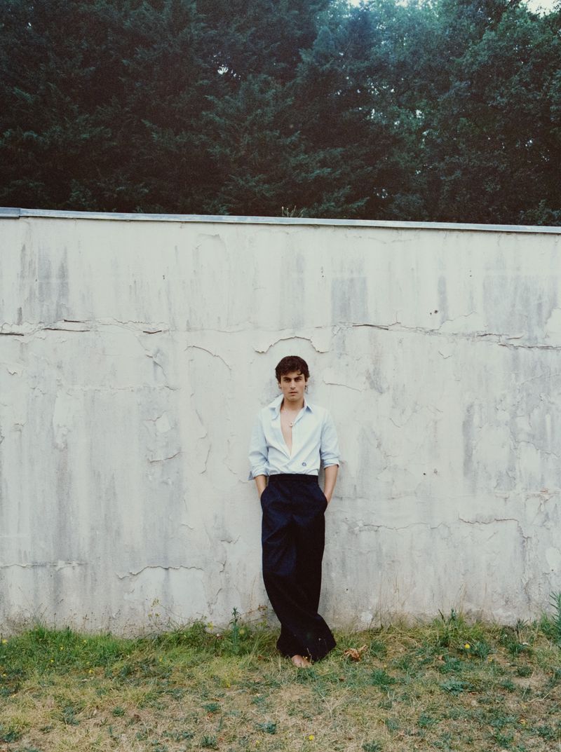Actor Julien de Saint Jean leaning barefoot against a cracked concrete wall wearing an unbuttoned blue shirt and black wide-leg trousers.