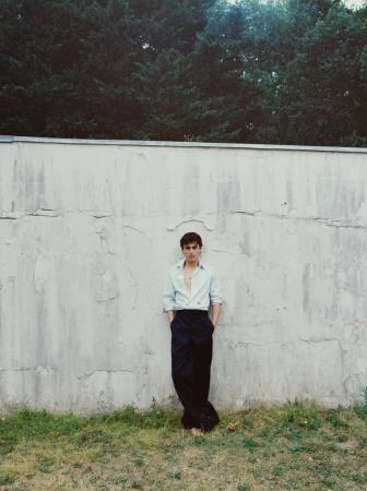 Actor Julien de Saint Jean leaning barefoot against a cracked concrete wall wearing an unbuttoned blue shirt and black wide-leg trousers.